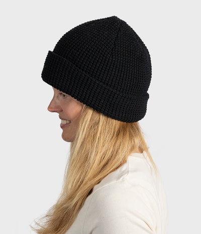 A woman in profile wearing a black ribbed knit beanie hat, shown against a plain light gray studio background.