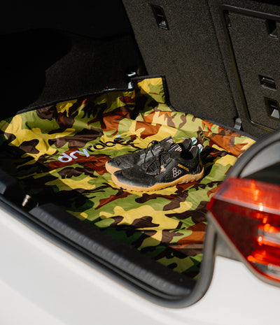 A pair of black athletic shoes placed on a camouflage-patterned floor mat inside a car trunk, with the brand name 'dryrobe' visible on the mat.