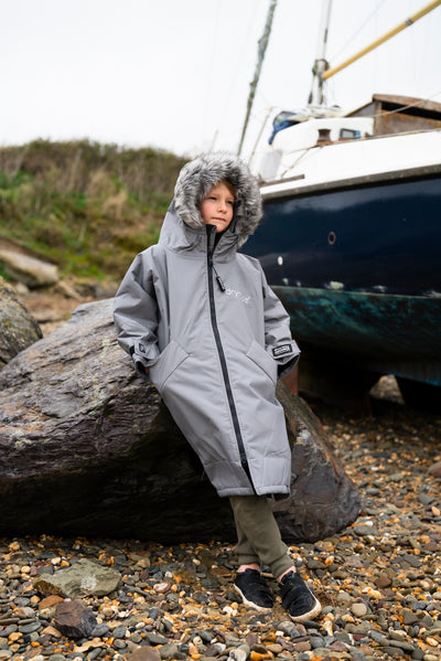 Child wearing a gray hooded coat standing on a rocky beach with a boat in the background.