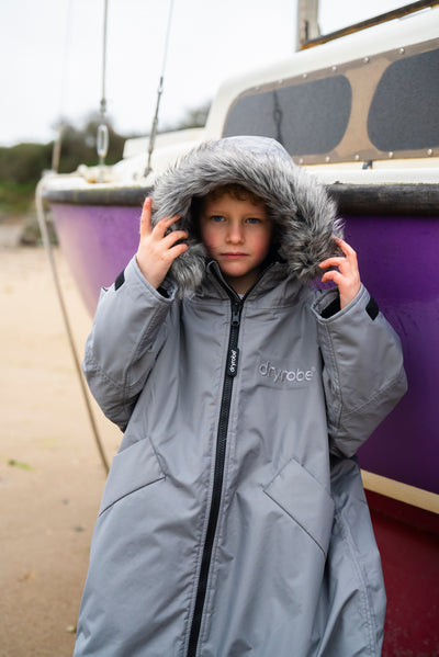 Child wearing a gray coat with a hood in front of a purple boat.