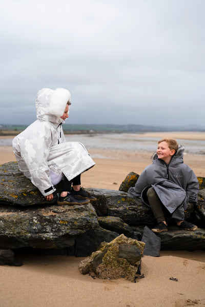 Two children in ponchos sitting on rocks by a beach on a cloudy day.