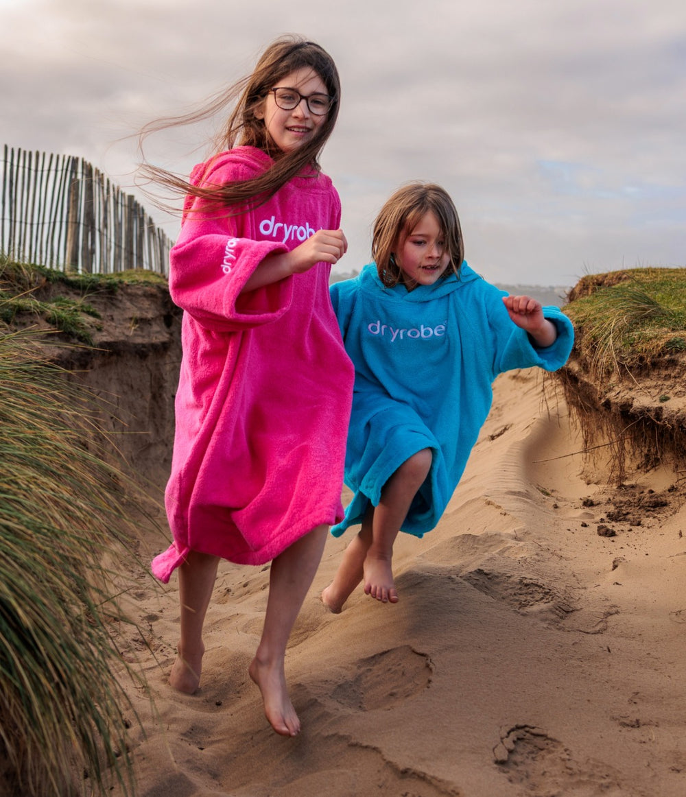 Two children running down a sandy beach dune wearing bright pink and blue Dryrobe, with dunes, grass, and an overcast sky in the background.