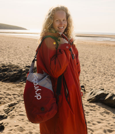 A woman standing on a sandy beach wearing a bright red waterproof Changing robe piece and carrying a red and gray Dryrobe compression bag. over her shoulder.