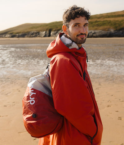 A man wearing a bright red waterproof Changing robe with a gray and white fleece-lined hood stands on a sandy beach, carrying a red and gray Dryrobe compression bag.