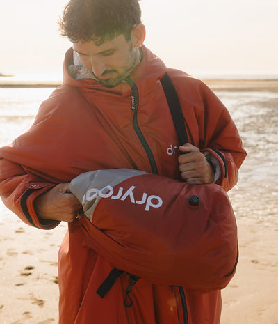 A man on a sandy beach wearing a rust-orange waterproof Dryrobe and holding a gray Dryrobe compression bag.