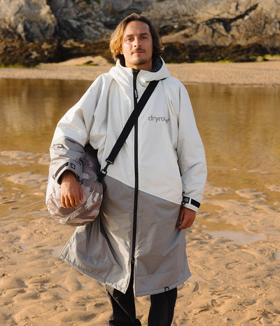 A man standing on a sandy beach wearing a long white and gray color-blocked waterproof changing robe with a Dryrobe compression bag.. The brand name 'dryrobe' is printed on the left chest.