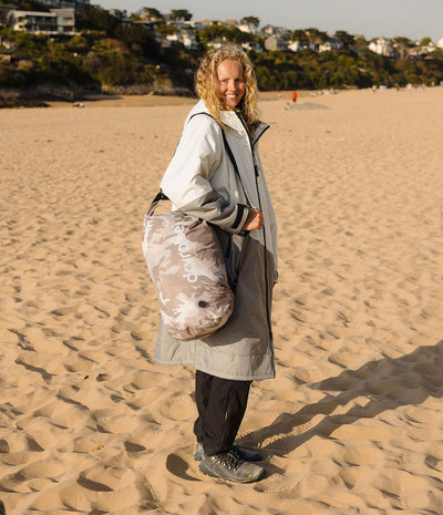 A woman standing on a sandy beach wearing a light gray hooded changing robe and carrying a large gray and camouflage-patterned Dryrobe compression bag. over her shoulder.