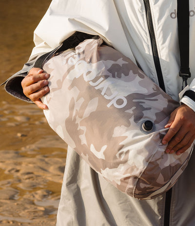 A person outdoors carrying a large Dryrobe compression bag. in a light beige and taupe camouflage cover with the brand name 'dlyrobe' printed in white. The person is wearing a white zip-up changing robe and the background shows a sandy, warm-toned outdoor ground.