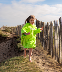 Child in a bright green towel robe running on a sandy path with wooden fencing and grassy area in the background.
