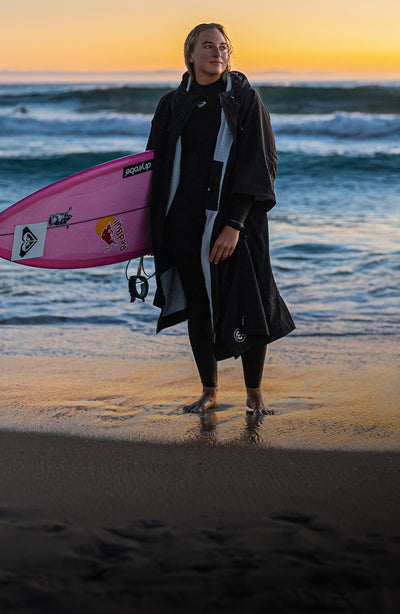 dryrobe® Ambassador Izzi Gomez stood on the beach holding surfboard, wearing dryrobe® Lite changing robe 