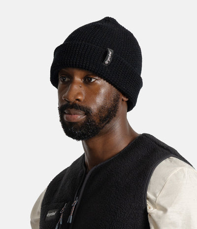 *MALE* A man wearing a black knit beanie with a small white 'dryrobe' brand tag and a black fleece vest over a cream shirt, photographed against a light gray studio background.