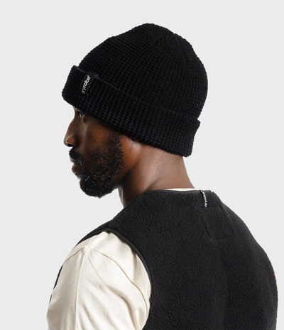 *MALE* A man in profile wearing a black ribbed knit beanie with a small white logo patch on the cuff, paired with a cream shirt and a black fleece vest, against a light gray studio background.