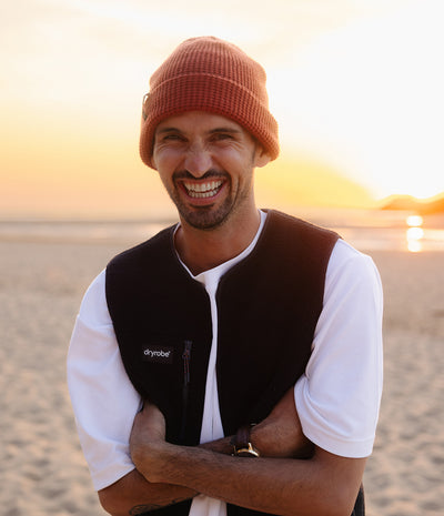 *MALE* A smiling man on a beach at sunset wearing a rust-orange knit beanie, a white t-shirt, and a black Dryobe fleece vest.