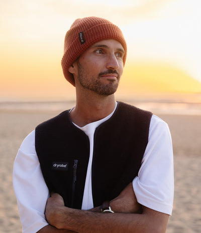 *MALE* A man wearing a rust-colored knit beanie and a black Dryrobe fleece vest over a white t-shirt, standing on a sandy beach at sunset.