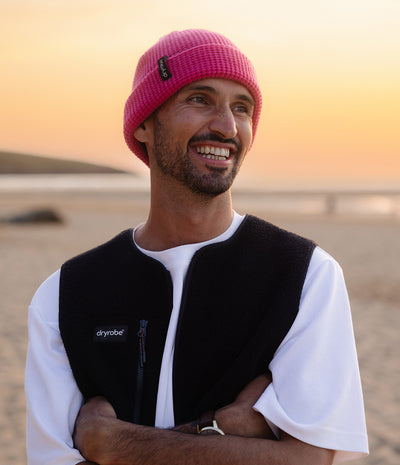 A smiling man on a beach at sunset wearing a bright pink knit beanie, a black Dryobe fleece vest over a white t-shirt, with his arms crossed.