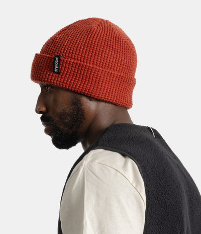 *MALE* A man in profile wearing a textured orange-red knit beanie with a black logo patch on the cuff, paired with a cream shirt and a dark fleece vest, against a light gray studio background.