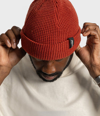 *MALE* A man wearing a rust-orange ribbed knit beanie hat with a small black brand tag, adjusting the brim with both hands against a light gray background.