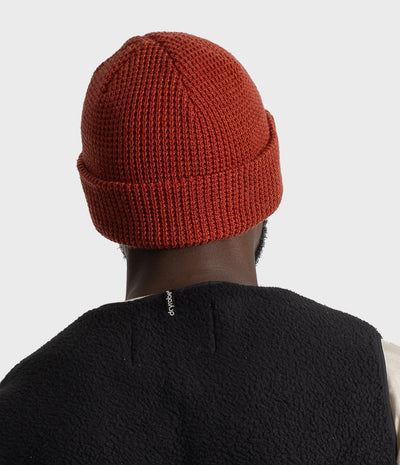 *MALE* A person shown from the back wearing a rust-orange ribbed knit beanie and a black fleece vest against a light gray studio background.
