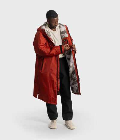 *MALE* A man wearing a long red hooded changing robe with a gray and black camouflage-patterned interior lining, styled with a white t-shirt, black pants, and beige sneakers, set against a plain light gray studio background.