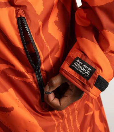 *MALE* Close-up of an orange patterned Dryrobe Changing Robe with a black zipper and a visible sleeve patch reading 'no-series ADVANCE PERFORMANCE'. A hand is shown zipping the jacket.