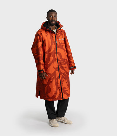 *MALE* A man modeling a long orange hooded Dryrobe Changing Robe with an abstract dark red-orange pattern, featuring a white 'swoosh' logo on the chest, black zippers, and black cuffs, paired with black pants and beige sneakers against a light gray background.
