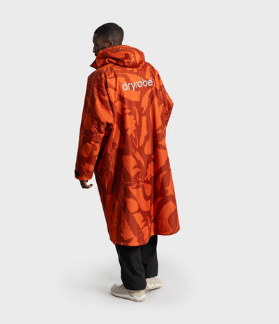 *MALE* A model shown from the back wearing a long orange hooded Dryrobe Changing Robe with a tonal abstract pattern and the brand name 'dryrobe' printed in white on the upper back, paired with black pants and light-colored sneakers against a light gray studio background.