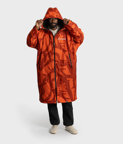 *MALE* A man wearing a long hooded Dryrobe Changing Robe with an orange and red abstract camouflage pattern, black pants, and beige sneakers, standing against a light gray studio background. The brand name 'dryear' is visible on the chest.