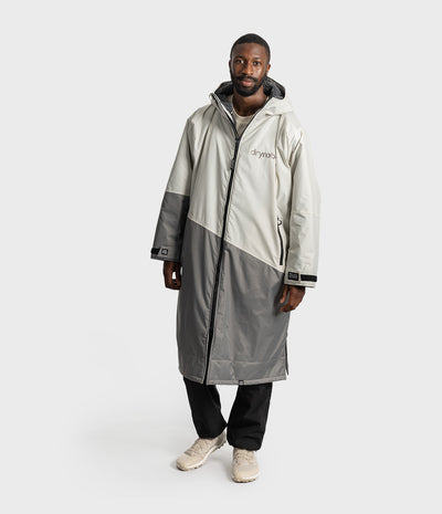 *MALE* A man wearing a Dryrobe, colour-blocked hooded in off-white and grey, with black pants and beige sneakers, standing against a light grey studio background.
