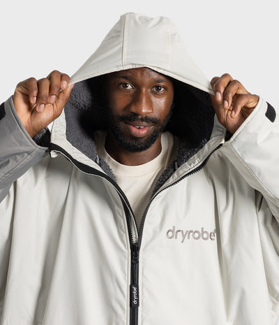 *MALE* A man wearing a light beige hooded Dryrobe holding the hood up with both hands against a clean white studio background.