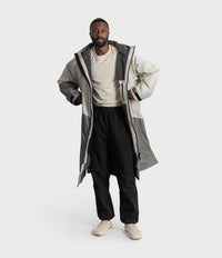 *MALE* A man modelling a long, oversized two-tone grey hooded Dryrobe over a cream t-shirt, black pants, and beige sneakers, set against a plain light grey studio background.