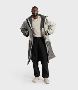 *MALE* A man modelling a long, oversized two-tone grey hooded Dryrobe over a cream t-shirt, black pants, and beige sneakers, set against a plain light grey studio background.
