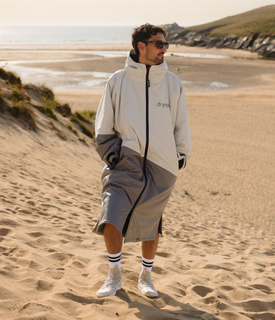 *MALE* A man wearing a light grey and dark grey split-colour hooded Dryrobe with a black zipper, standing on a sandy beach dune with the ocean and cliffs in the background.
