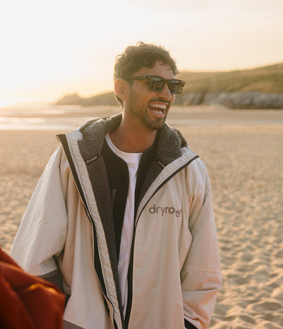 *MALE* A man wearing a beige Dryrobe waterproof jacket with a dark grey sherpa lining, black sunglasses, and a white t-shirt, standing on a sandy beach during golden hour.