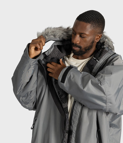 *MALE* A man wearing a gray hooded dryrobe jacket with a gray faux-fur trimmed hood and a dark gray fleece lining, shown from the chest up against a light gray studio background.
