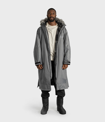 *MALE* A man modeling a long gray hooded Dryrobe with a faux-fur trimmed hood, worn over a cream sweatshirt, black pants, and black boots, against a light gray studio background.