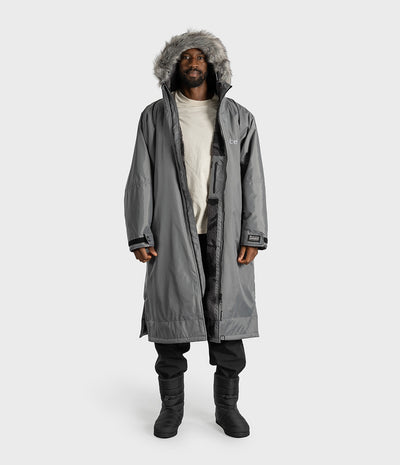 *MALE* A man wearing a long gray dryrobe with a gray faux-fur trimmed hood, layered over a cream t-shirt, black pants, and black boots, standing against a light gray studio background.