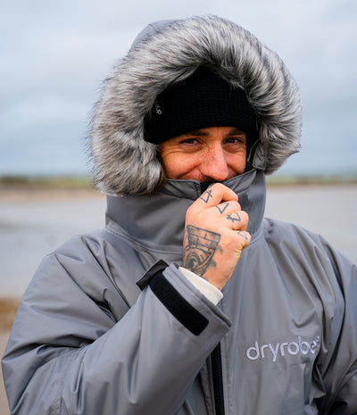 *MALE* A man wearing a gray Dryrobe hooded jacket with a gray faux-fur trimmed hood, a black knit beanie, and a white shirt, standing outdoors against an overcast sky.