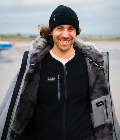 *MALE* A smiling man wearing a black beanie and a black zip-up fleece over a black Dryrobe with gray camouflage panels, gray faux-fur trim, and a white undershirt, standing outdoors on a beach with an overcast sky.