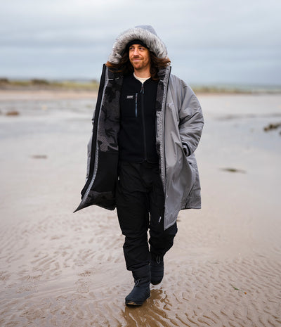 *MALE* A man walking on a sandy beach wearing a long gray hooded Dryrobe with a black and gray patterned lining, a black zip-up hoodie, black pants, and black boots.