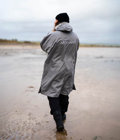 *MALE* A person viewed from behind walking on a sandy beach wearing a long gray hooded Dryrobe with 'dryrobe' printed on the back, black pants, and a black beanie.
