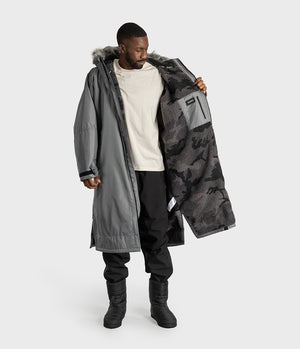 *MALE* A man modeling a long gray parka with a black and gray camouflage patterned interior lining, shown against a light gray studio background.