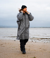 A man wearing a long gray waterproof Dryrobe, black beanie, and black pants walks along a sandy beach under an overcast sky.