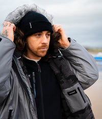 A man wearing a gray outdoor Dryrobe with a gray faux-fur hood trim, a black knit beanie with a small white logo, and a black zip-up fleece layer, standing outdoors against an overcast sky.