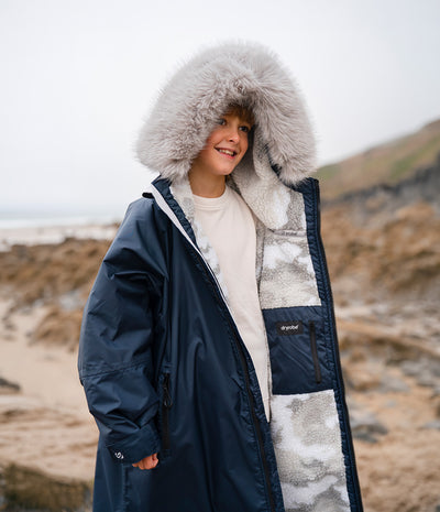 *MALE* A child wearing a navy blue waterproof Dryrobe with a large fluffy light gray fur-trimmed hood and a light gray sherpa-lined front, standing outdoors on a rocky beach.