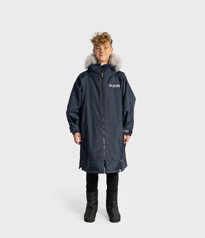*MALE* A child model wearing a navy blue Dryrobe kids' with a light gray faux-fur hood trim, black pants, and black boots, standing against a light gray studio background.