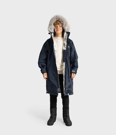 *MALE* A child model wearing a navy blue hooded Dryrobe with a light gray faux-fur trim, layered over a cream sweater, black pants, and black boots, set against a plain light gray studio background.