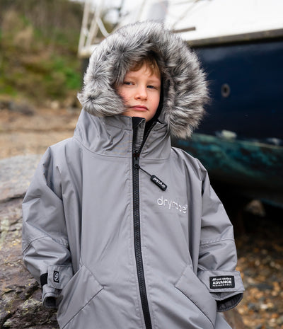 *MALE* A child wearing a gray Dryrobe waterproof jacket with a faux-fur trimmed hood, standing outdoors in front of a boat.