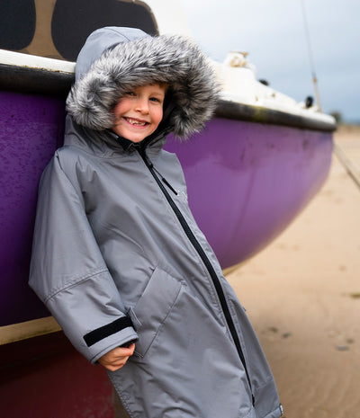 *MALE* A smiling child wearing a gray hooded Dryrobe with a faux-fur trimmed hood, standing beside a purple boat on a sandy beach.