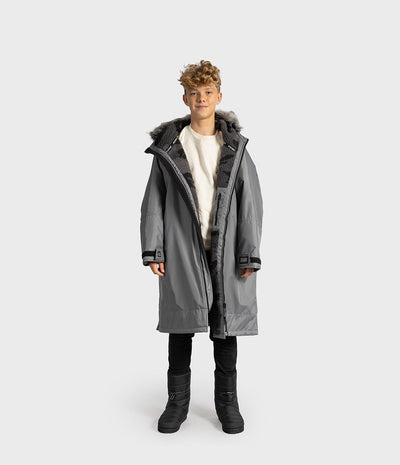 *MALE* A child model wearing a long gray hooded Dryrobe with dark faux-fur trim, a cream shirt, black pants, and black boots, standing against a light gray studio background.