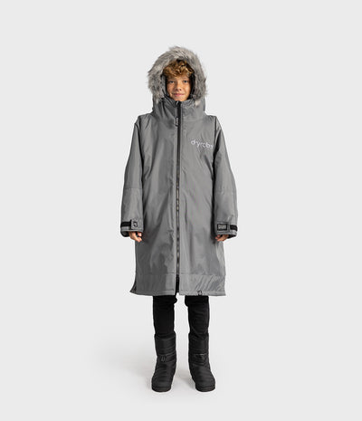 *MALE* A child wearing a medium gray hooded Dryrobe with faux-fur trim, black pants, and black boots, standing against a light gray studio background. The brand name 'dyrobe' is printed on the left chest.